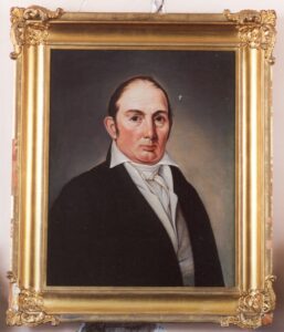 19th century painting of a white man with short red hair, dark eyes, a high collar, and black jacket against a grey backdrop. The painting is in a gold frame.